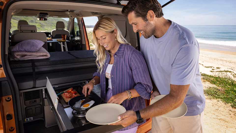 Junges Paar brät Spiegeleier in der Heckküche des VW California am Strand