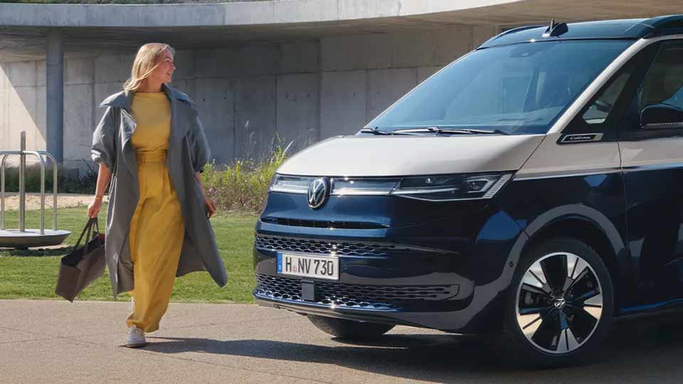 Junge Frau steht neben einem weiß-blauen VW California